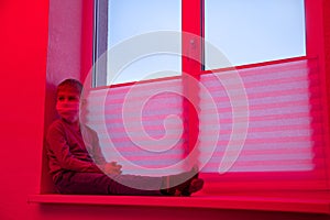 Sad boy in danger sitting on windowsill in protective mask