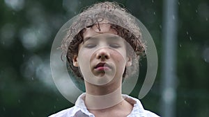 A sad boy gets wet in the rain. Psychological problems of teenagers, outdoor portrait of a curly teenager in the summer