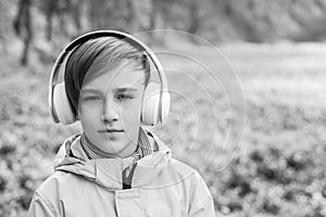 Sad boy listening to music at nature. Kid with headphones relaxing in the spring park. Upset boy with headphones on the walk