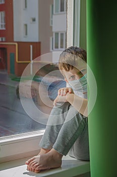 Sad boy in a medical protective mask wants to walk on the street, a child sits on the windowsill and yearns. Quarantine self-