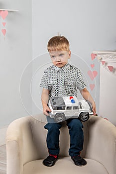 A sad boy sits on the back of a chair and holds in his hands a large toy car. Childhood concept