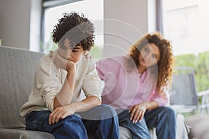 Sad boy sitting on sofa in living room with his mother in background