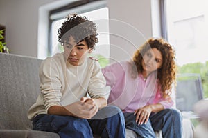 Sad boy sitting on sofa in living room with his mother in background