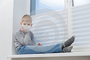 Sad boy sitting on windowsill in protective mask