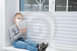 Sad boy sitting on windowsill in protective mask holding heart