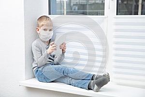 Sad boy sitting on windowsill in protective mask holding tablet