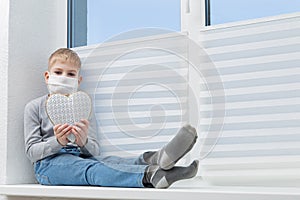 Sad boy sitting on windowsill in protective mask holding white heart