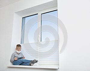 Sad boy sitting on windowsill in protective mask playing game on tablet