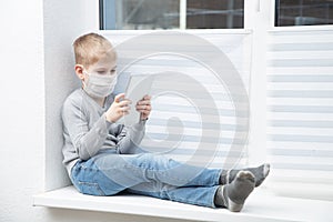 Sad boy sitting on windowsill in protective mask playing tablet