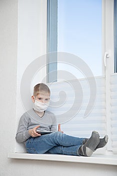 Sad boy sitting on windowsill in protective mask with tablet