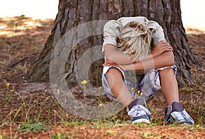 Sad boy, stress and depression by tree with anxiety, mental health or bored on grass in nature. Male person, child or