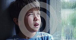 Sad boy, window and bored on a rainy day, lonely and looking outside on a wet winter day. Child, depression and thinking