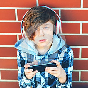 Sad boy with wireless headphones playing mobile games outside school. Technology, internet addiction