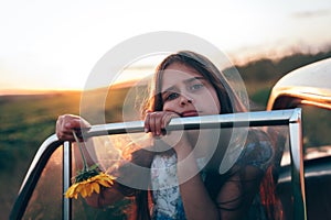 Sad brunette teen girl with sunflower in hand leaned on the car door and looking at the camera, sunset on the background