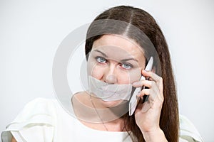 Sad brunette woman with cellphone and tape on her lips, grey background