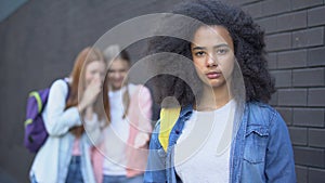 Sad bullying victim looking camera, female classmates teasing african schoolgirl