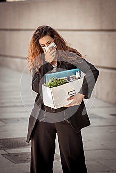 Sad business woman with face mask and box of office staff being fired due to coronavirus job cuts