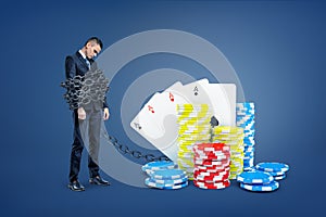 A sad businessman bound by a large metal chain to pile of casino cards and colorful chips.