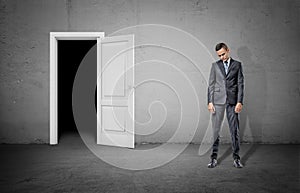 A sad businessman with his head low stands near a door frame showing complete darkness.