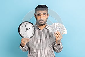 Sad businessman holding big fan of dollar banknotes and wall clock, time is money, looking at camera