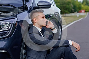 Sad businessman use smartphone sitting on road near the broken car opened the hood, calling car assistance services,