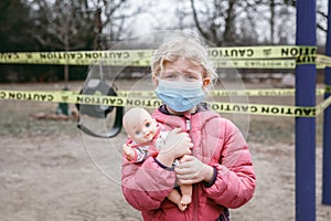 Sad Caucasian girl in face mask with baby toy on closed playground outdoor. Kids play area locked with yellow caution tape in