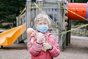 Sad Caucasian girl in face mask with baby toy on closed playground outdoor. Kids play area locked with yellow caution tape in