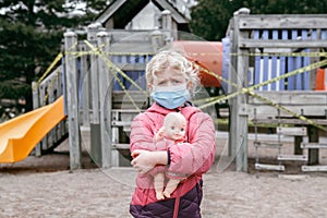 Sad Caucasian girl in face mask with baby toy on closed playground outdoor. Kids play area locked with yellow caution tape in