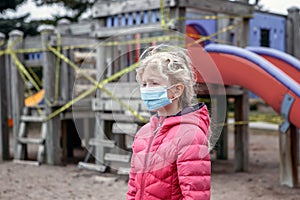 Sad Caucasian girl in face mask standing on closed playground outdoor. Kids play area locked with yellow caution tape in Toronto,