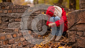 Sad caucasian girl in a red coat and beret sits on a brick wall on a walk in autumn.