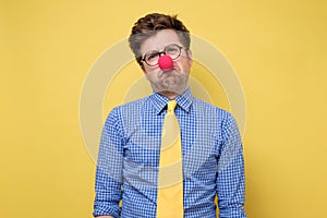 Sad caucasian man wearing clown red nose on yellow wall.