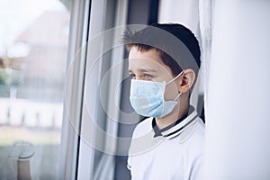 Sad child boy in medical mask looking through the window. Quarantine