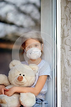 Sad Child, little girl in mask with teddy bear looking from window, coronavirus quarantine