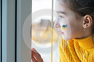 A sad child look at the window with the flag of Ukraine painted on the cheek, worries and fear. Humanitarian aid to children,