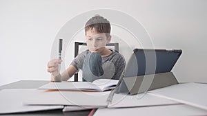 Sad child sitting at desk with tablet computer and many books. Boy feels bored, because he do not understand homework