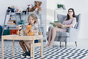 A sad child with trauma playing with toys and a professional psychologist sitting in an armchair in the background