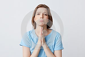 Sad concerned young woman in blue t shirt with hands folded in praying position have problems and begging for help isolated over