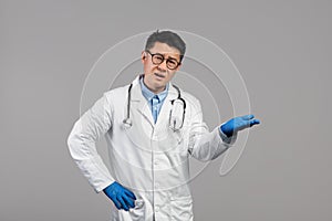 Sad confused adult asian man doctor in white coat, goggles and protective gloves holds empty space on his hand