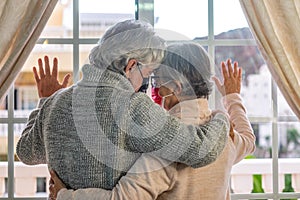 A sad couple of senior people hugging each other in front of the window while staying at home for coronavirus infection
