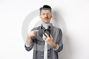 Sad crying guy pointing at smartphone and sobbing, complaining or feeling miserable, standing on white background