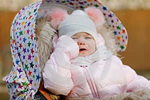 Sad crying hungry baby girl sitting in the pram or stroller on cold autumn, winter or spring day. Weeping child in warm
