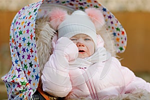 Sad crying hungry baby girl sitting in the pram or stroller on cold autumn, winter or spring day. Weeping child in warm