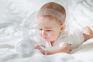 Sad and crying Infant baby girl lying on the white bed with special bottle of water with nipple. Tries to gnaw her. teething