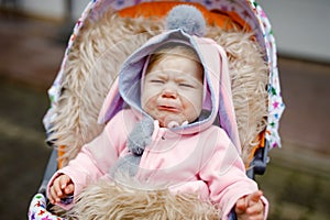 Sad crying little beautiful baby girl sitting in the pram or stroller on autumn day. Unhappy tired and exhausted child