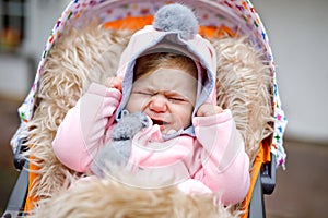 Sad crying little beautiful baby girl sitting in the pram or stroller on autumn day. Unhappy tired and exhausted child