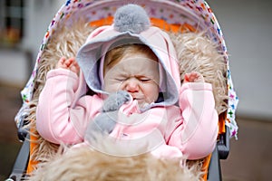 Sad crying little beautiful baby girl sitting in the pram or stroller on autumn day. Unhappy tired and exhausted child