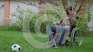 Sad crying military man in wheelchair looking at soccer ball on green summer meadow on backyard. Wide shot of depressed