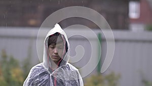 Sad depressed boy standing in a raincoat in the rain, slow motion