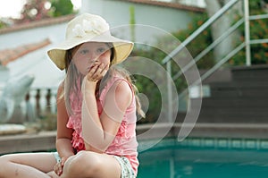 A sad, depressed little girl in a hat sitting near a pool