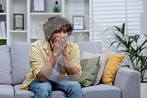 Sad depressed man sitting alone at home on sofa in living room, desperate indian man